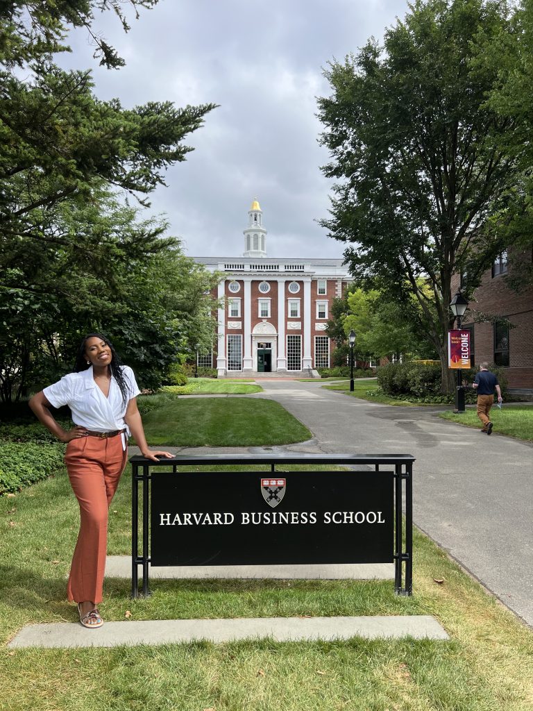 Photo of a Black woman in a white shirt and brown slacks posing next to a sign for Harvard Business School
