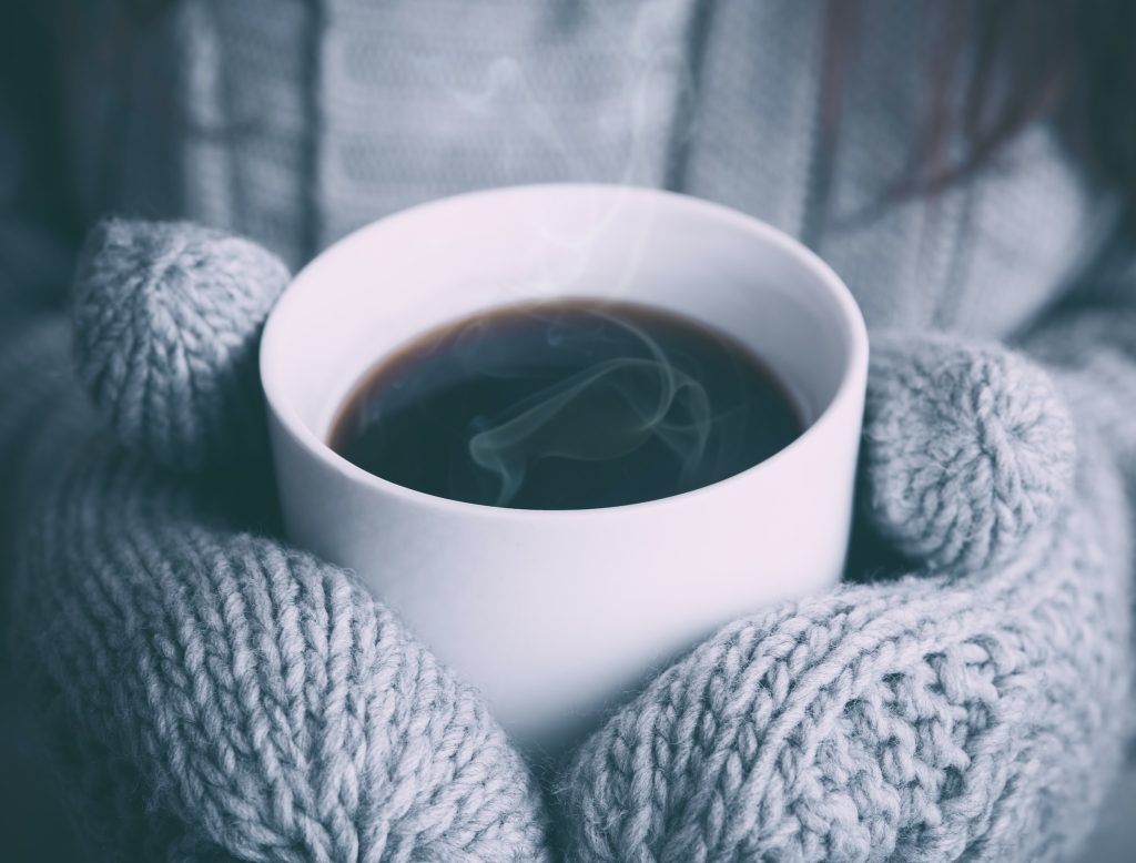 Hands covered in light grey, knit mittens clutch a steaming mug of coffee. Image by Alex Padurariu on Unsplash.