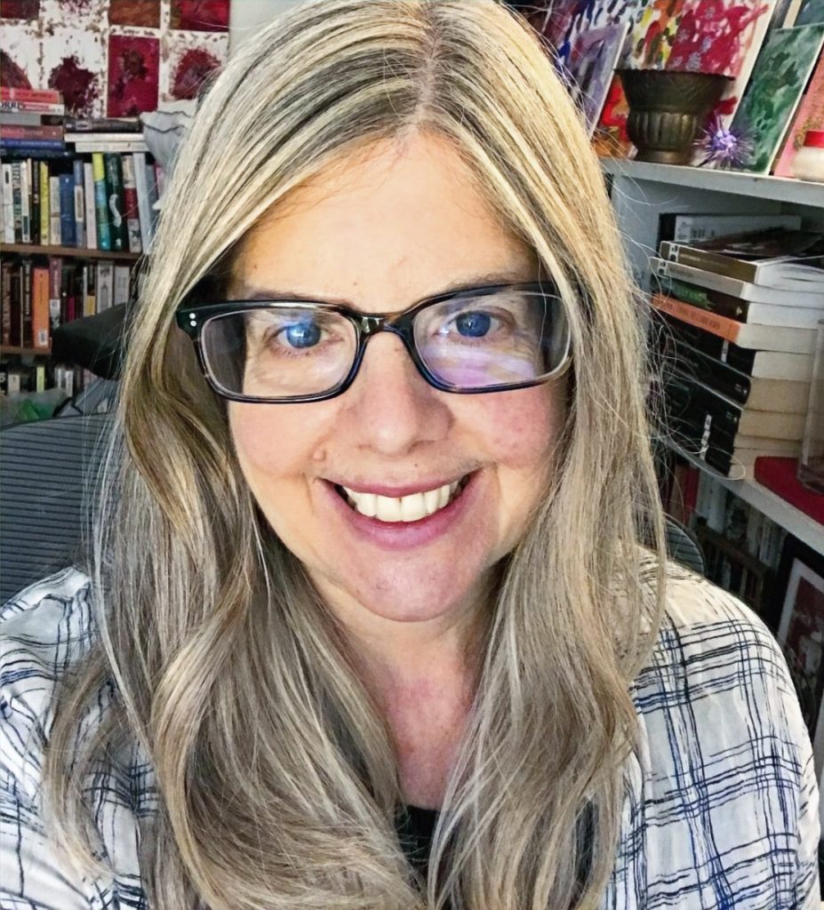 Headshot of Lorraine Martindale with long blonde hair, wearing brown eyeglasses and a white top with black linear design, in front of bookshelves