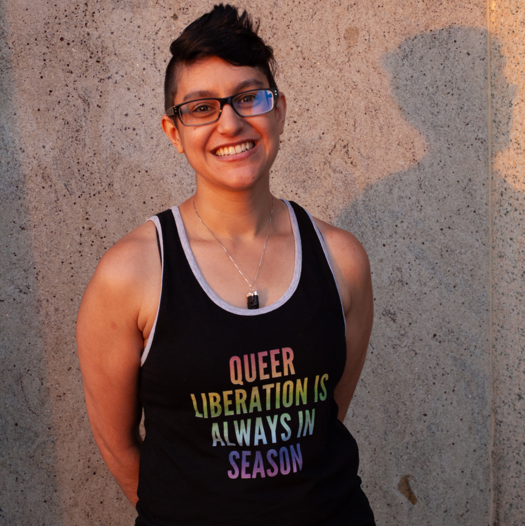 Headshot in front of a beige speckled wall of Dominique Chatterjee, a South Asian (Tamil/Bengali) Dutch-American person with black cropped hair, wearing a sleeveless top imprinted with the rainbow-color words "Queer liberation is always in season"