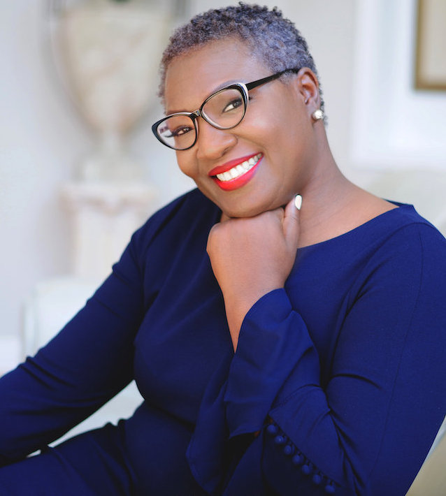Smiling Black woman with glasses and a blue dress, sitting with her hand propping up her chin