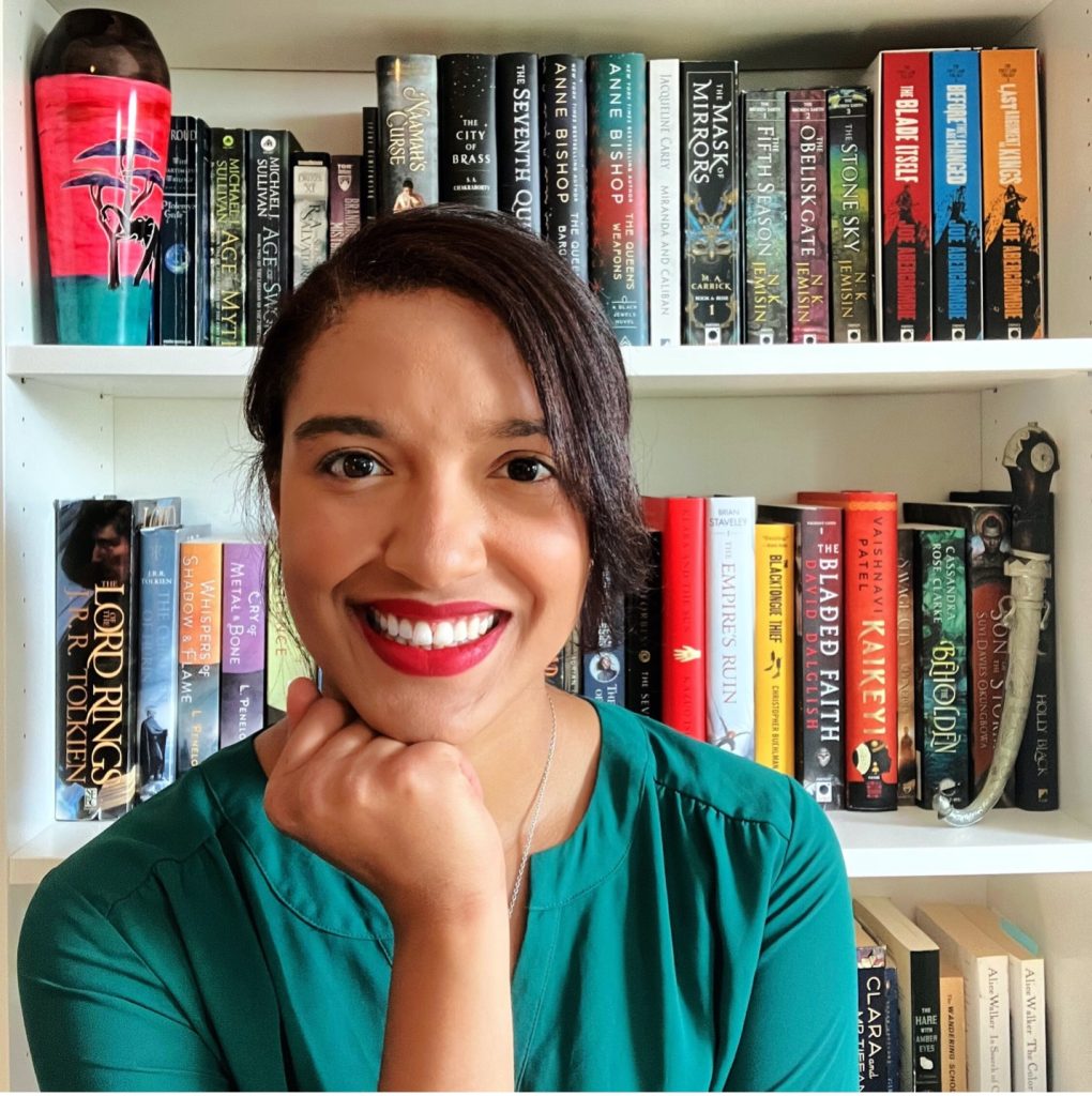 Smiling Black woman in front of a bookshelf, wearing a teal top and propping up her chin with with her hand.
