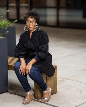Sharese Terrell Willis in a black top, jeans, and sandals sitting on a bench in front of a building